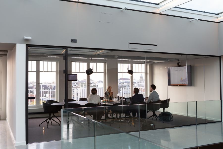 Photo of the outside of a conference room with glass windows, a team meeting inside, and a video screen at the front of the room.