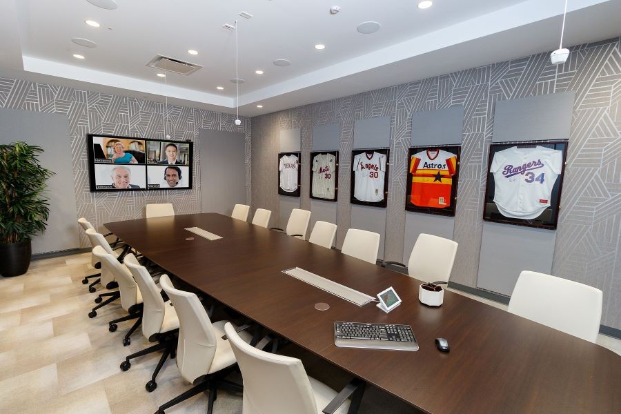 A video conference room with in-ceiling speakers, microphones, a control panel, and four people on the display.