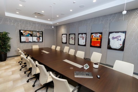 A video conference room with in-ceiling speakers, microphones, a control panel, and four people on the display.